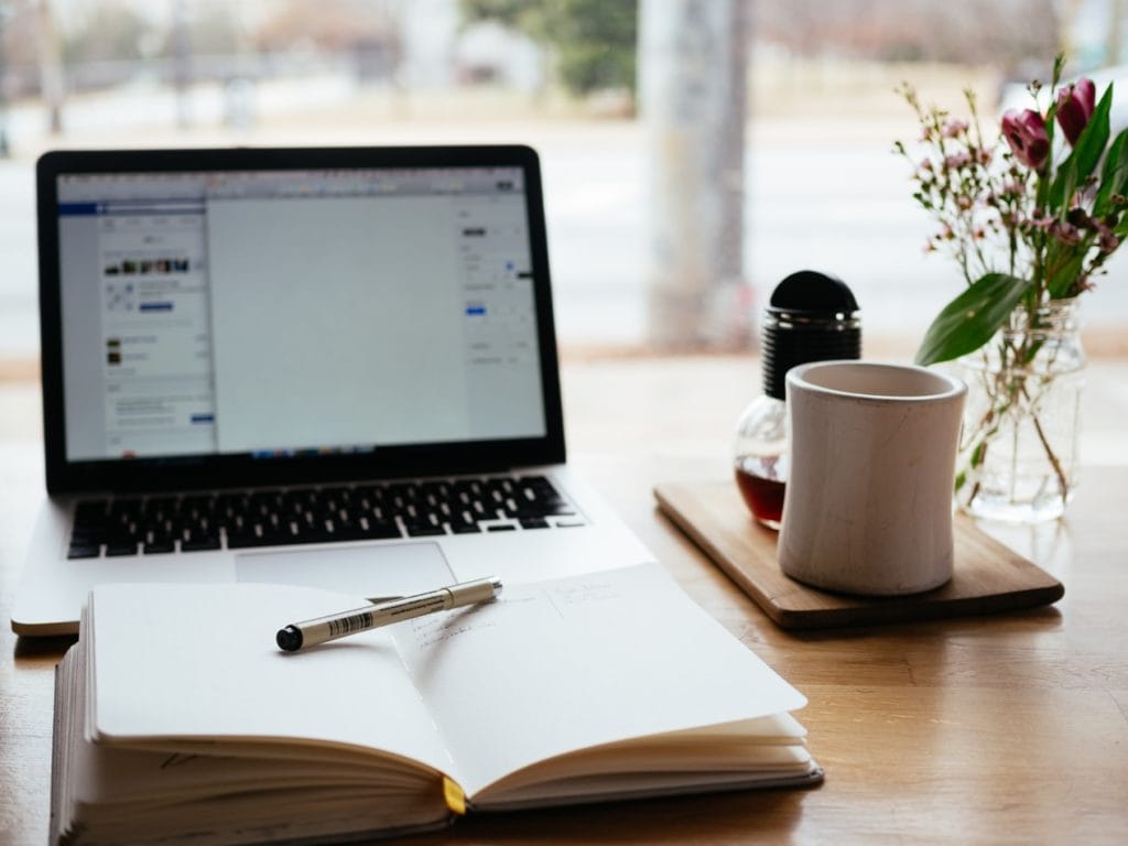 Image of a laptop, notepad and pen, mug and flowers on a table.