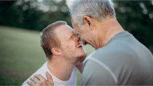 Two men with their heads pressed together, laughing.  One of the men has learning disabilities. 